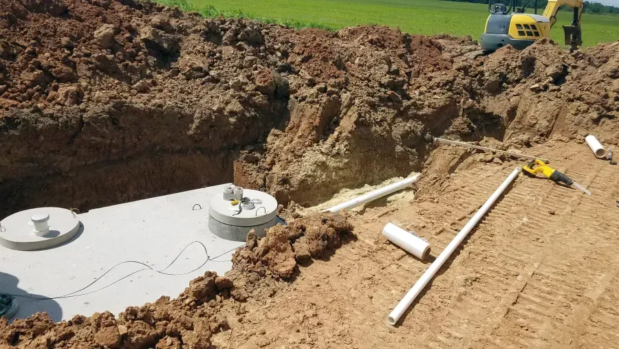 Excavation pit with exposed concrete tanks, white pipes, and a yellow digger on a dirt construction site.