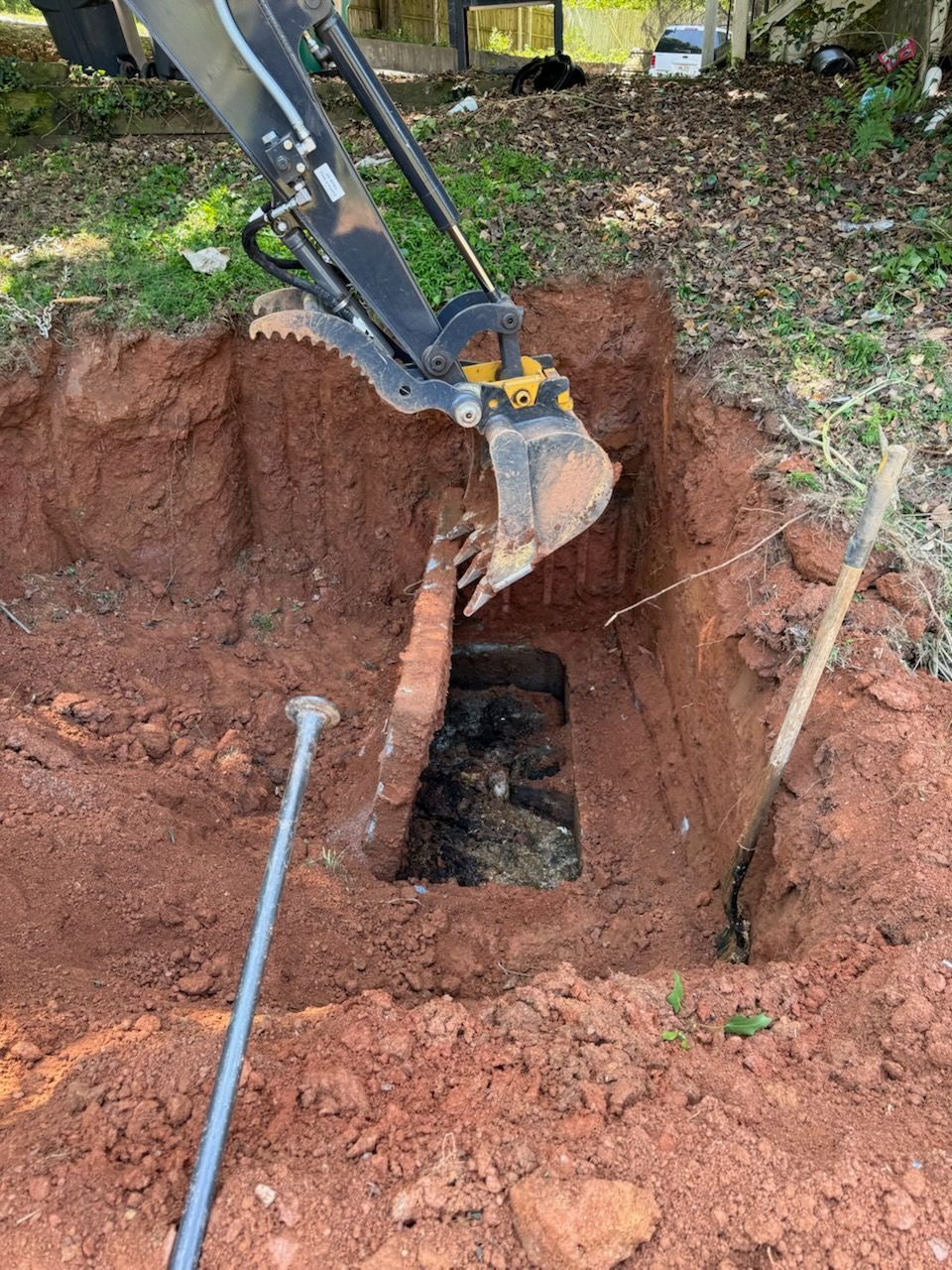 Excavator digging a deep rectangular trench in reddish soil beside a yard with tools nearby