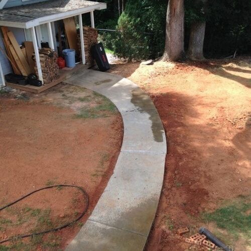 Curved concrete walkway through a dirt yard beside a house and trees