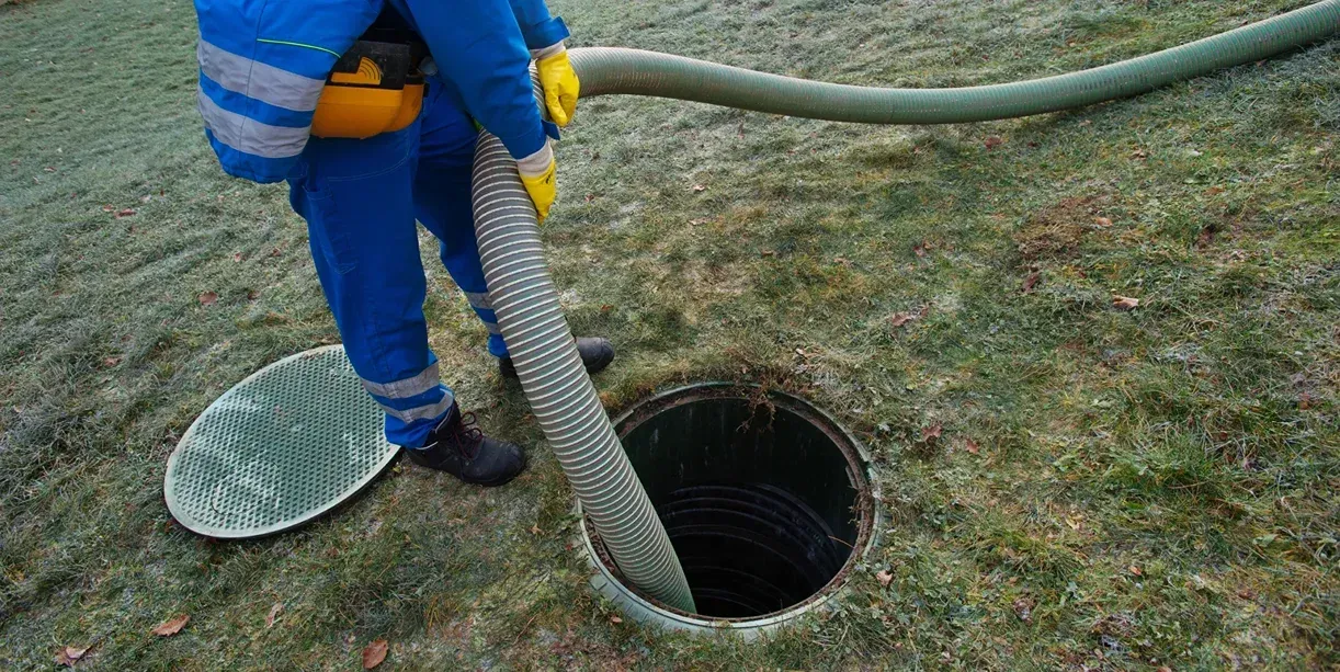 Worker in blue uniform using a hose to inspect a dark circular manhole on grass