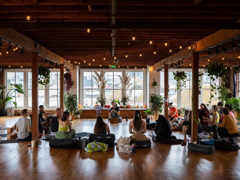People seated on meditation cushions in a bright studio with large windows and hanging lights