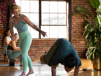 Yoga class in a bright studio, instructor helping a bent-over participant near a large window