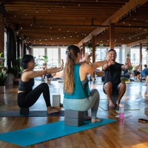 People doing a yoga class on mats in a bright wooden studio, arms raised in seated poses