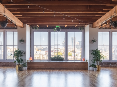 Empty loft studio with large windows, exposed beams, polished wood floor, and city view.