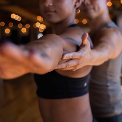 Two people stretching with arms extended in a dim studio, warm lights blurred in the background