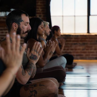 People seated in a yoga class, meditating with hands pressed together in a sunlit studio