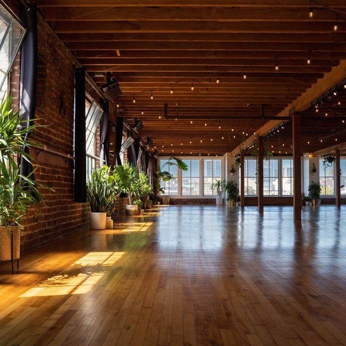Sunlit loft with exposed brick, wooden beams, plants, and polished floors overlooking water