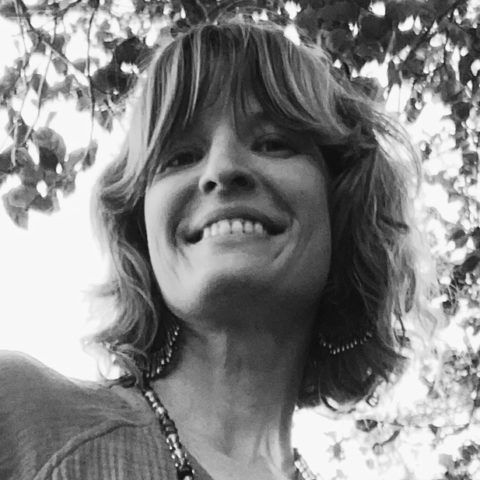 Smiling person with shaggy hair, seen from below against leafy trees