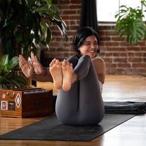 Person doing a yoga boat pose on a mat in a bright studio, smiling toward the camera.