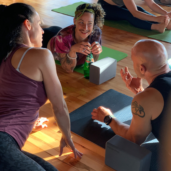 People seated on yoga mats in a sunlit studio, chatting and smiling with yoga blocks nearby