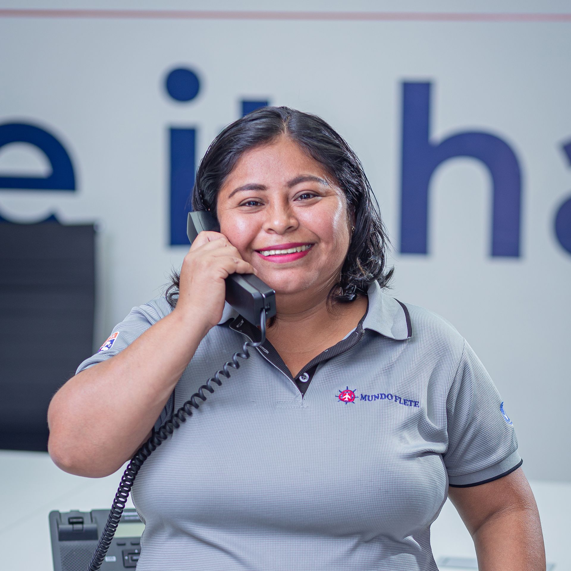 Persona sonriente con un polo gris sosteniendo un auricular de teléfono en un entorno de oficina.
