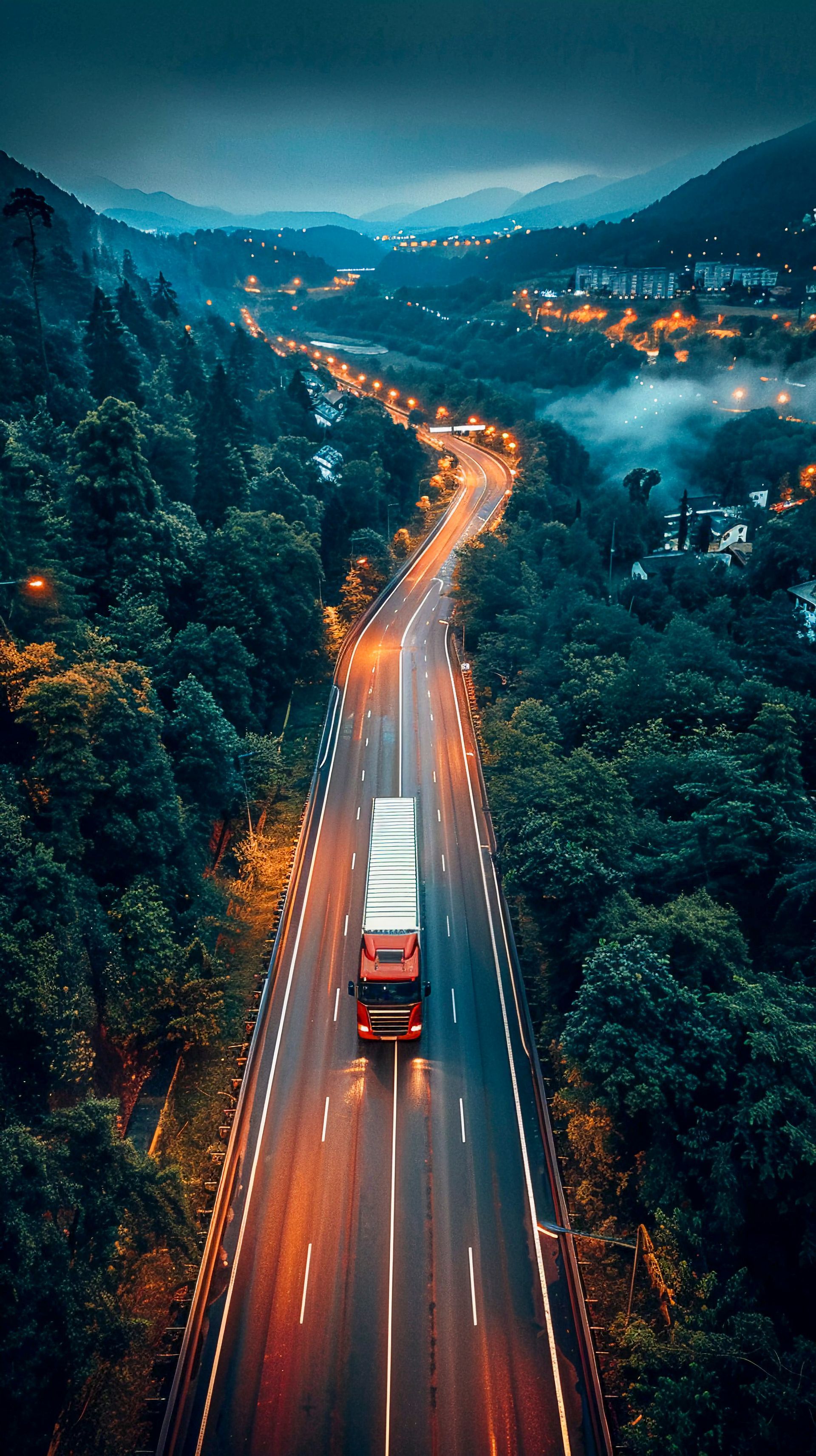 Una sinuosa carretera de montaña al atardecer, con un camión rojo y las luces brillantes de la ciudad entre colinas boscosas y envueltas en la niebla.