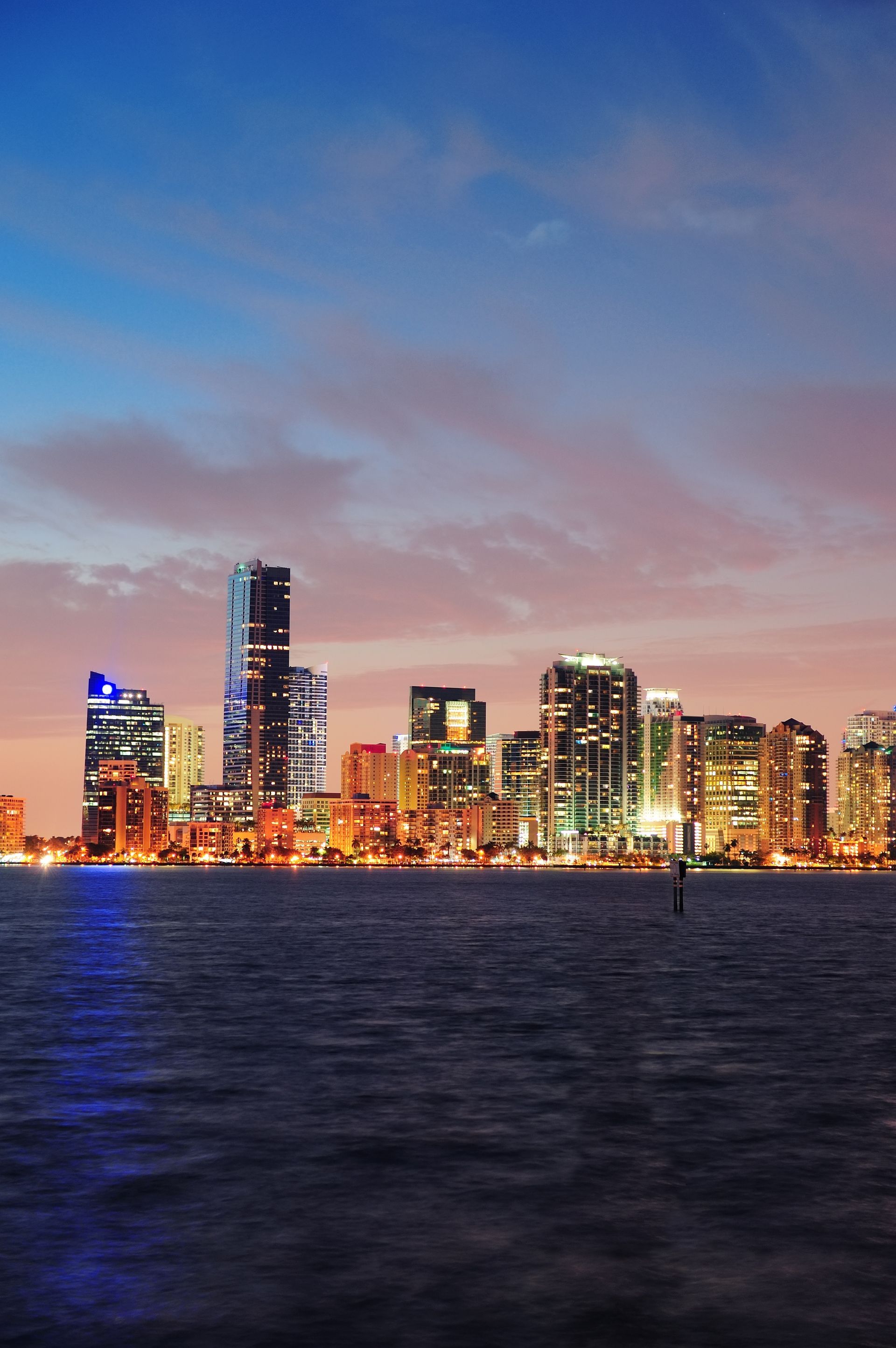 El horizonte de la ciudad al atardecer se refleja en el agua oscura, con edificios resplandecientes bajo un cielo rosa azulado.