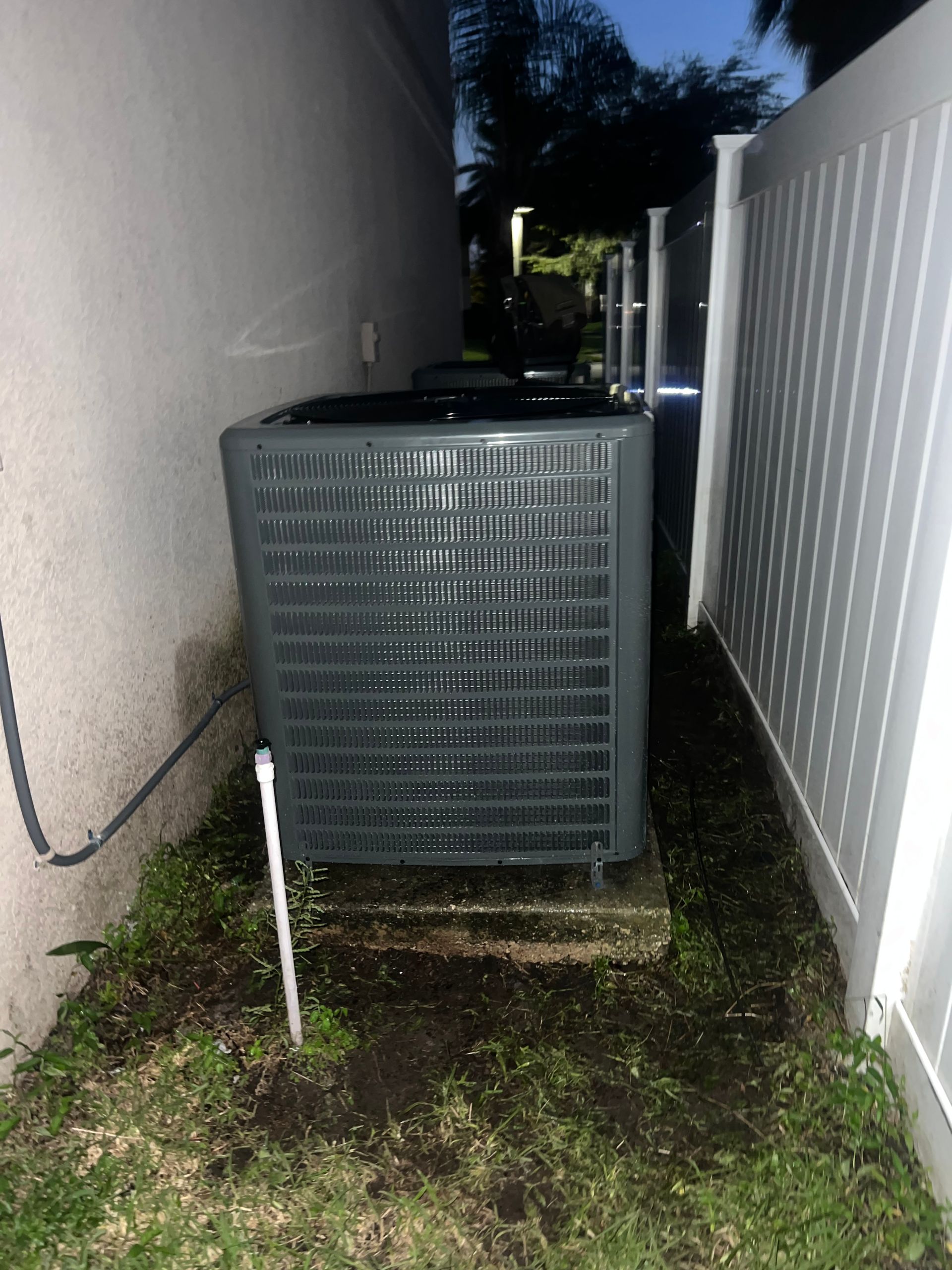 a large air conditioner is sitting in the backyard next to a white fence .