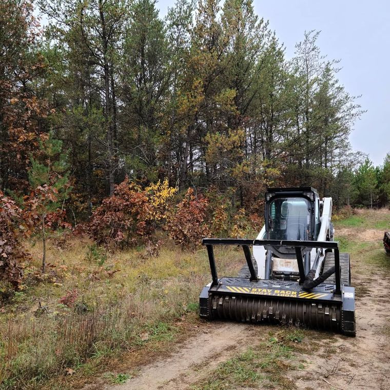 Compact track loader clearing a dirt path beside autumn trees and brush