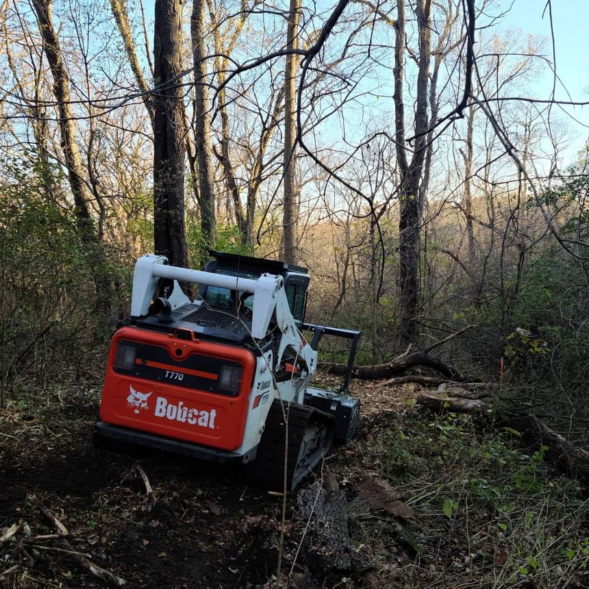 Red Boissat skid-steer loader in a wooded area, clearing brush on muddy ground.