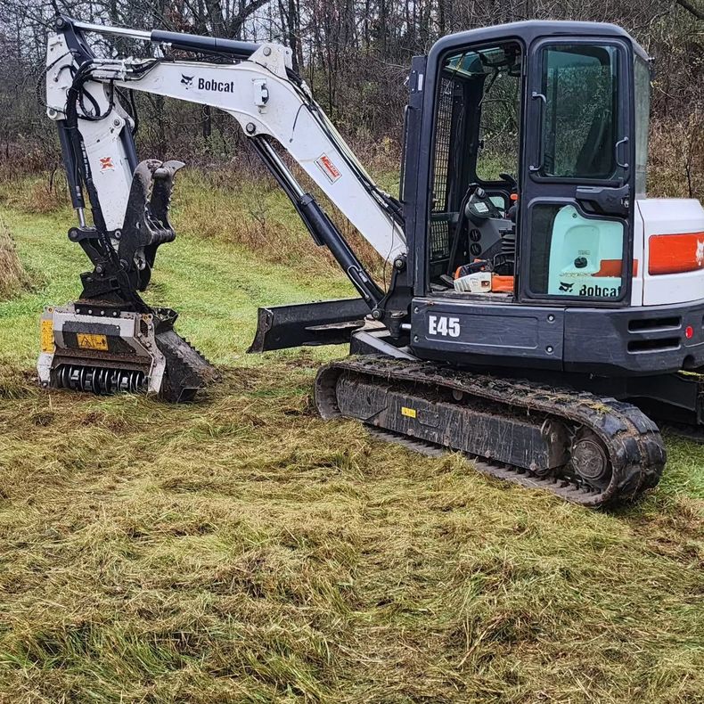 Compact excavator clearing grass on a muddy field, using a mowing attachment.