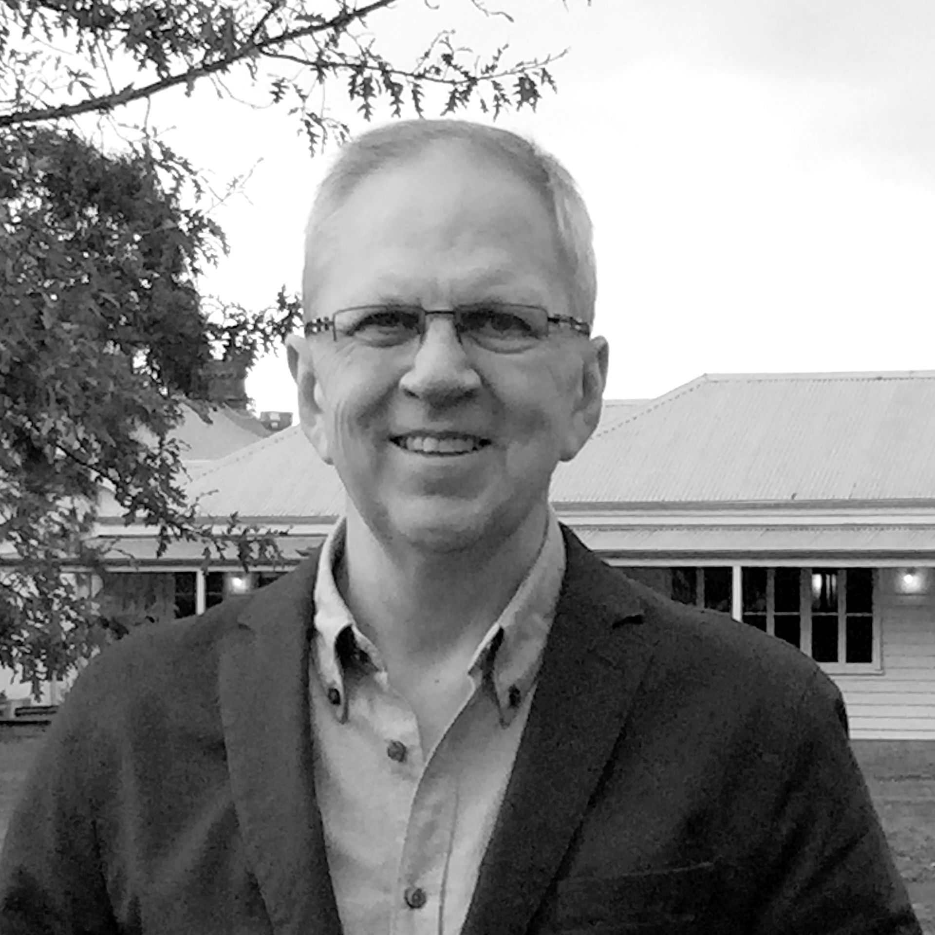 A man in a suit and glasses is smiling in front of a house.