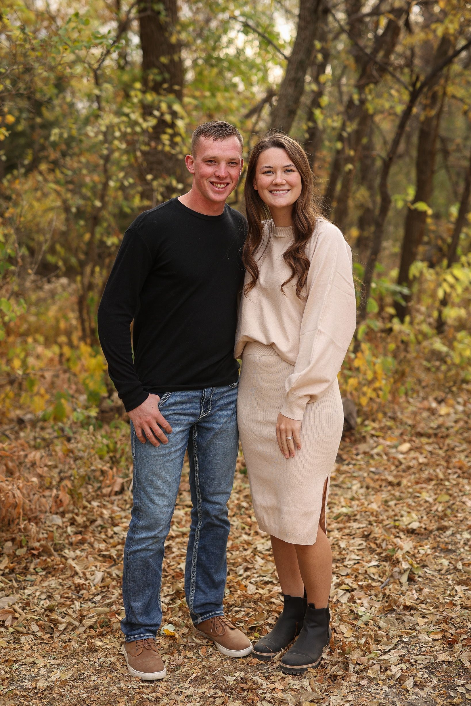 Couple in fall attire poses in wooded area. Man in black sweater and jeans, woman in beige knit outfit.
