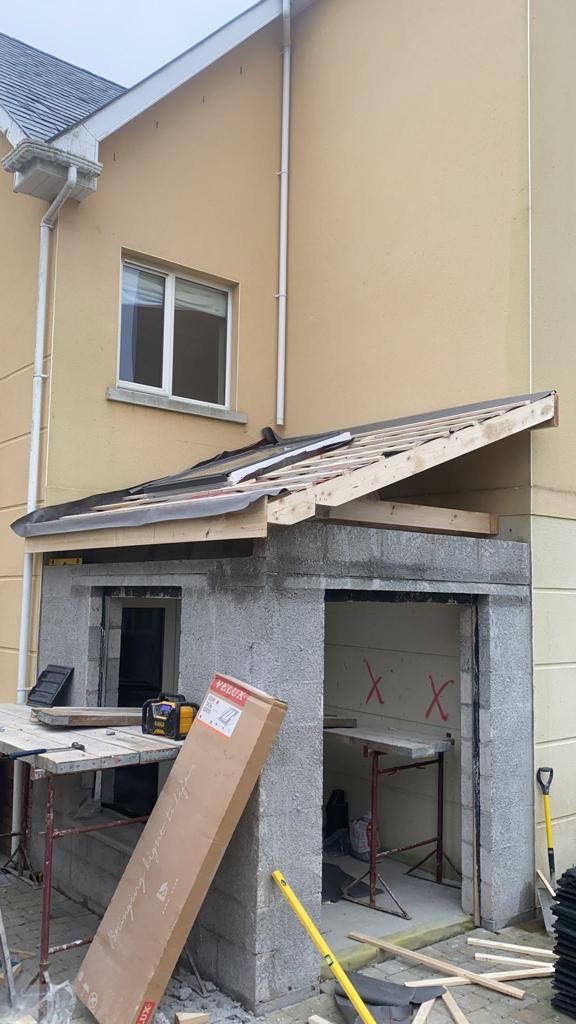 Construction of a lean-to roof addition next to a two-story beige house. Partially built concrete block structure.