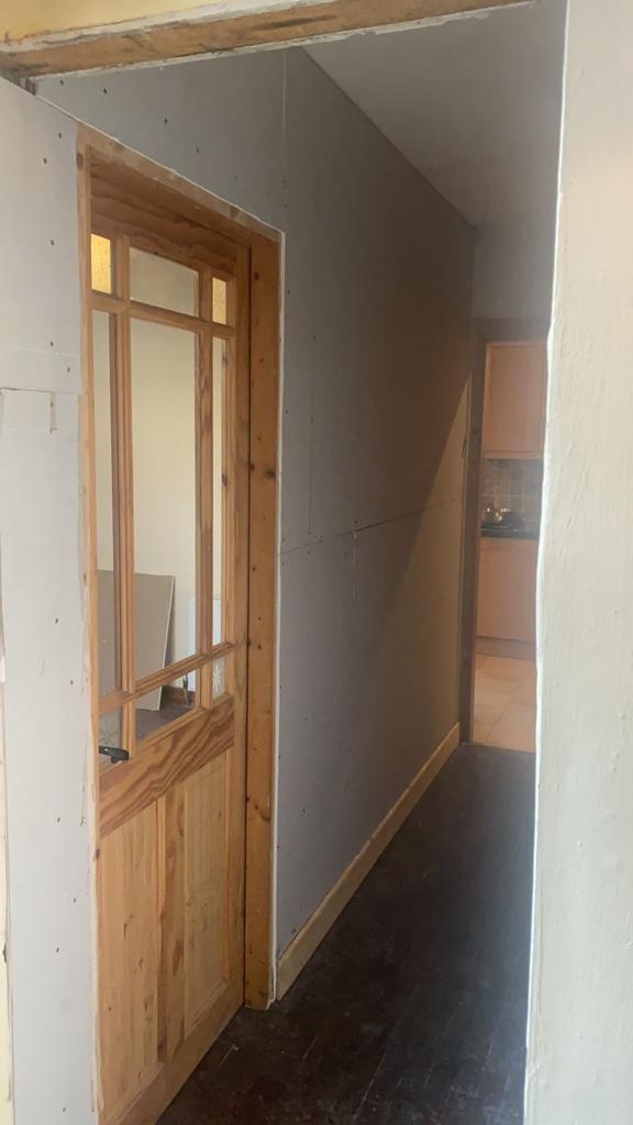 Hallway with wooden door on the left, gray wall, and dark floor. Light streams from doorway.