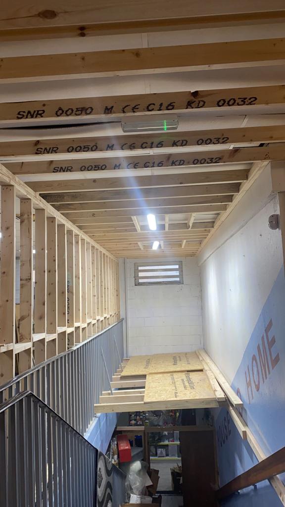 Stairwell under construction; wooden framing, plywood, and exposed beams. Fluorescent lights illuminate the space.