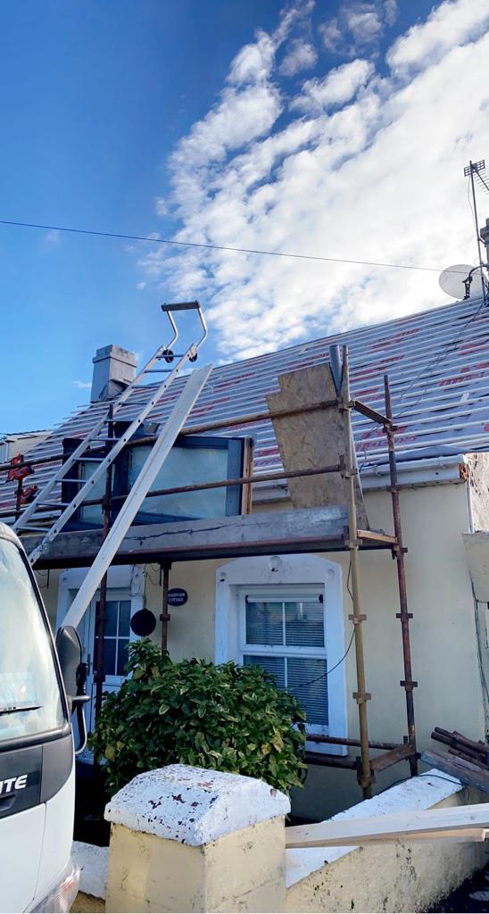 House undergoing roof work with scaffolding. Partly cloudy sky, light yellow building, red/gray roof tiles.