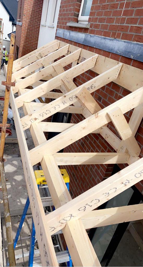 Wooden roof structure being built against a red brick building; scaffolding and a ladder are visible.