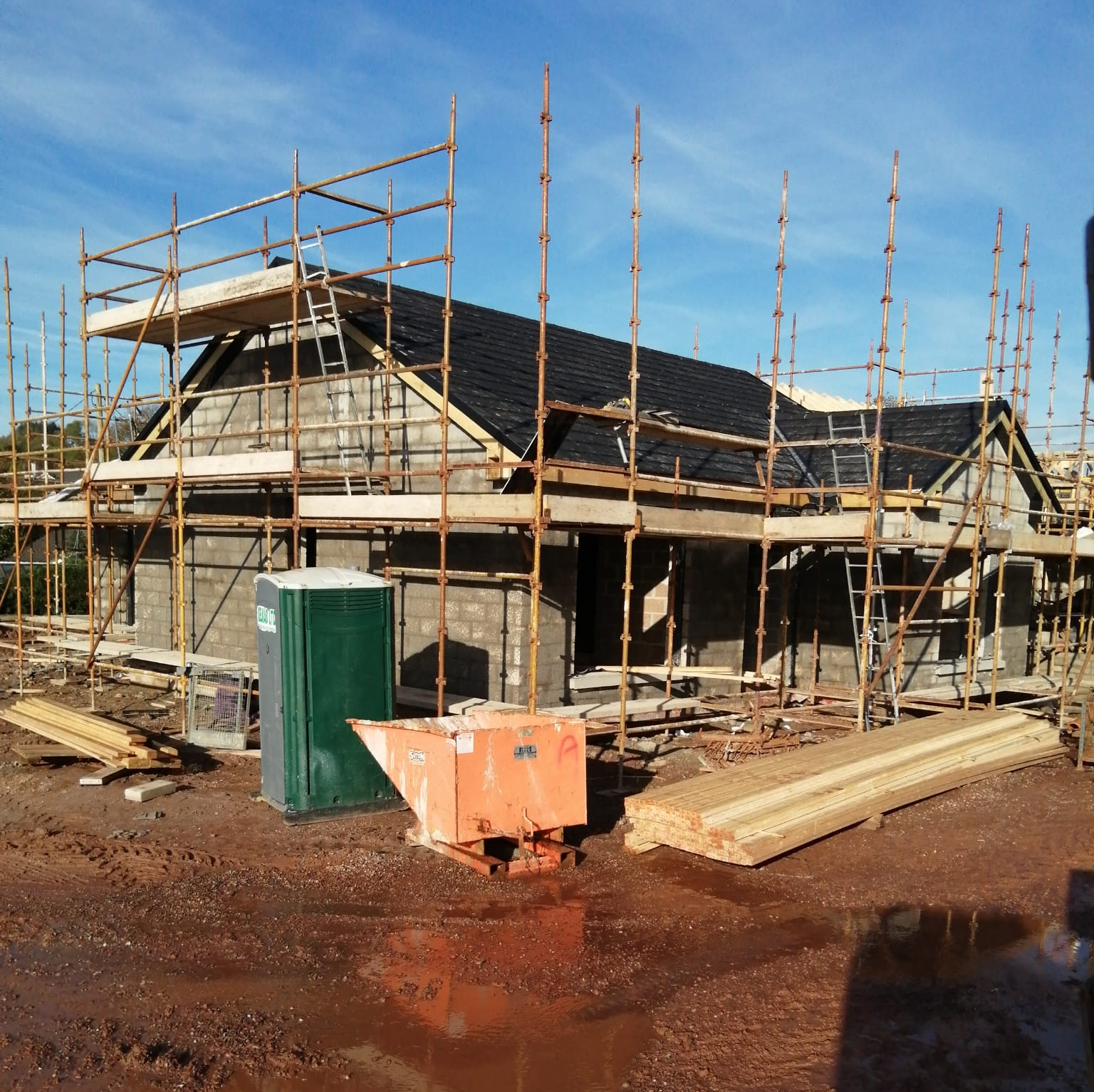 House under construction with scaffolding, portable toilet, and concrete mixer on a muddy site.