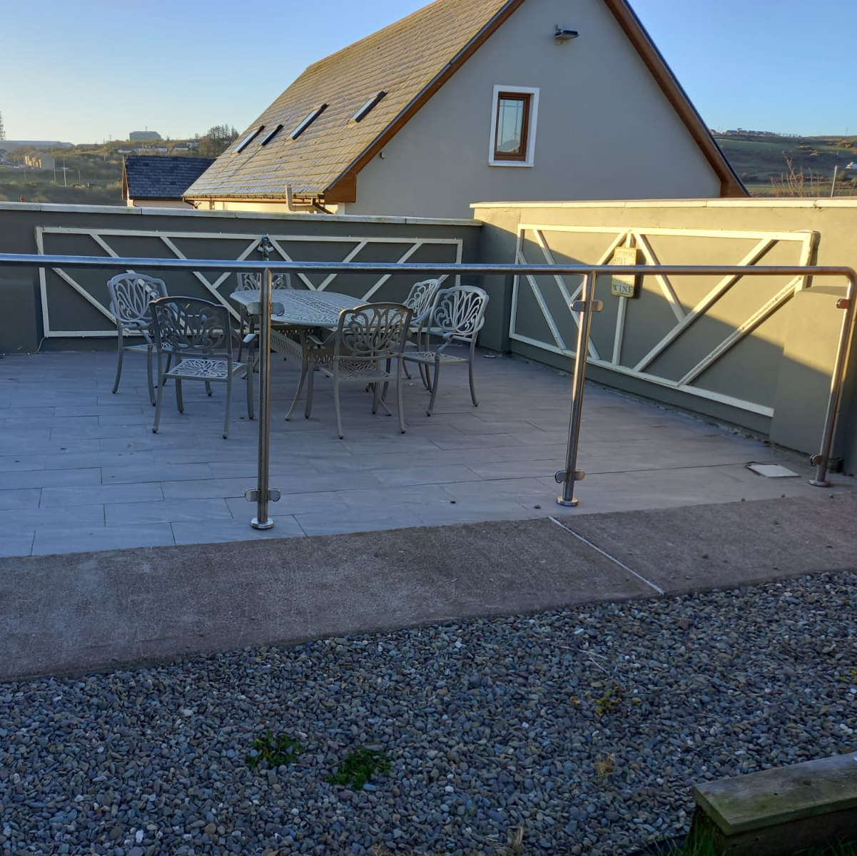 Outdoor patio with gray stone, metal railing, table, chairs, and light-colored house in background.