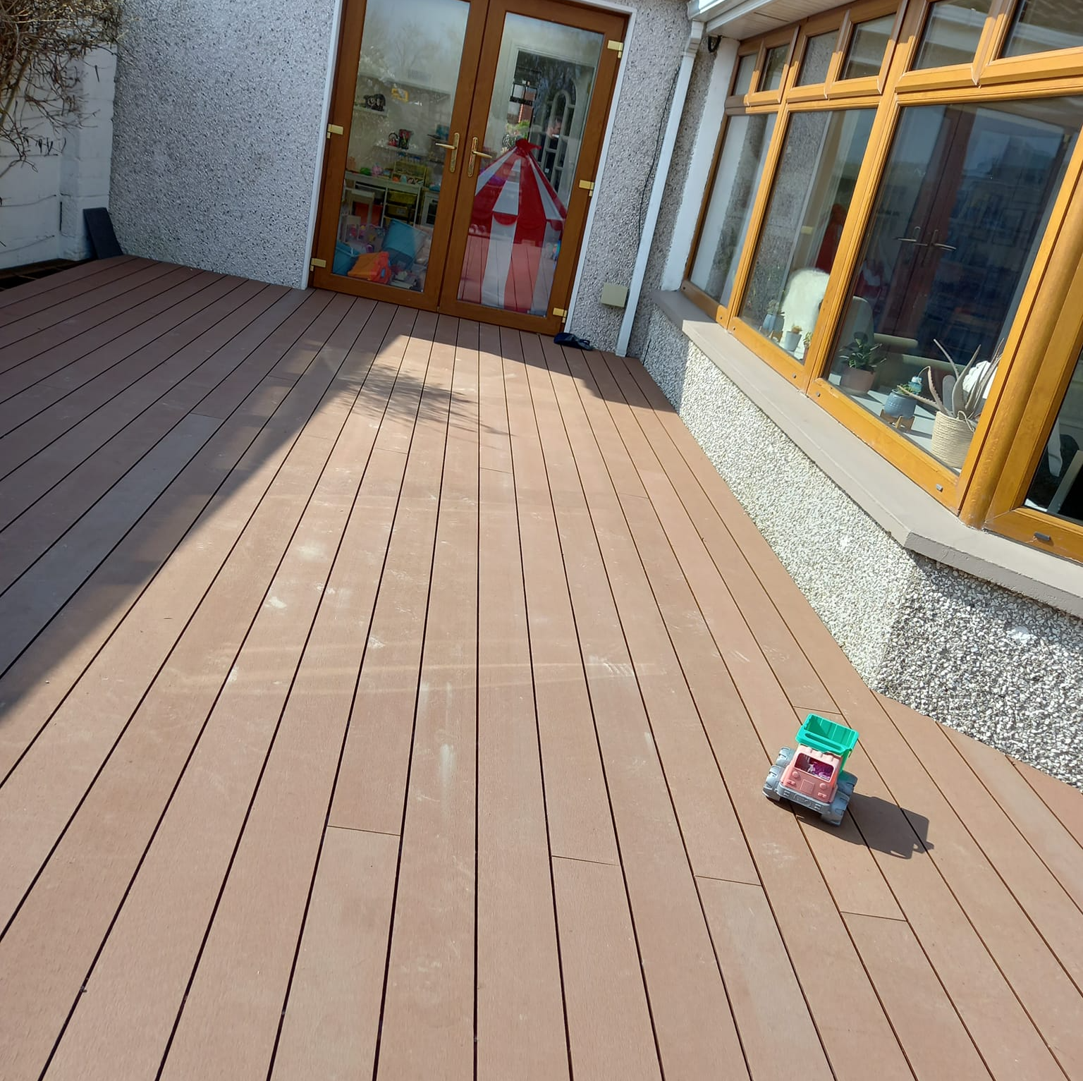 Wooden deck outside a house with glass doors and windows. A toy truck sits on the deck.