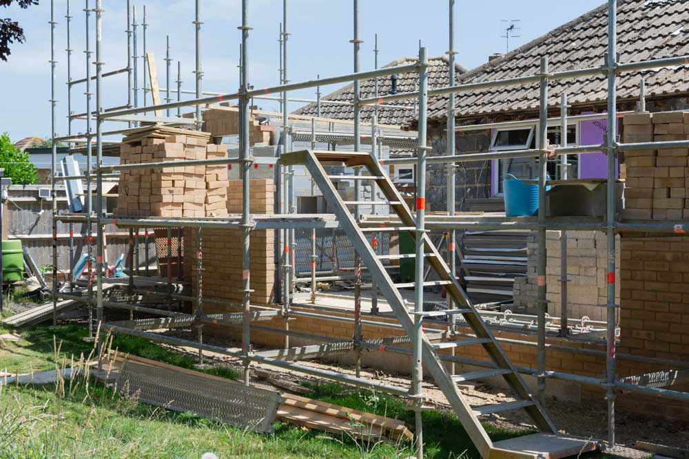 Scaffolding surrounds a building under construction, stacks of bricks visible, sunny outdoors.