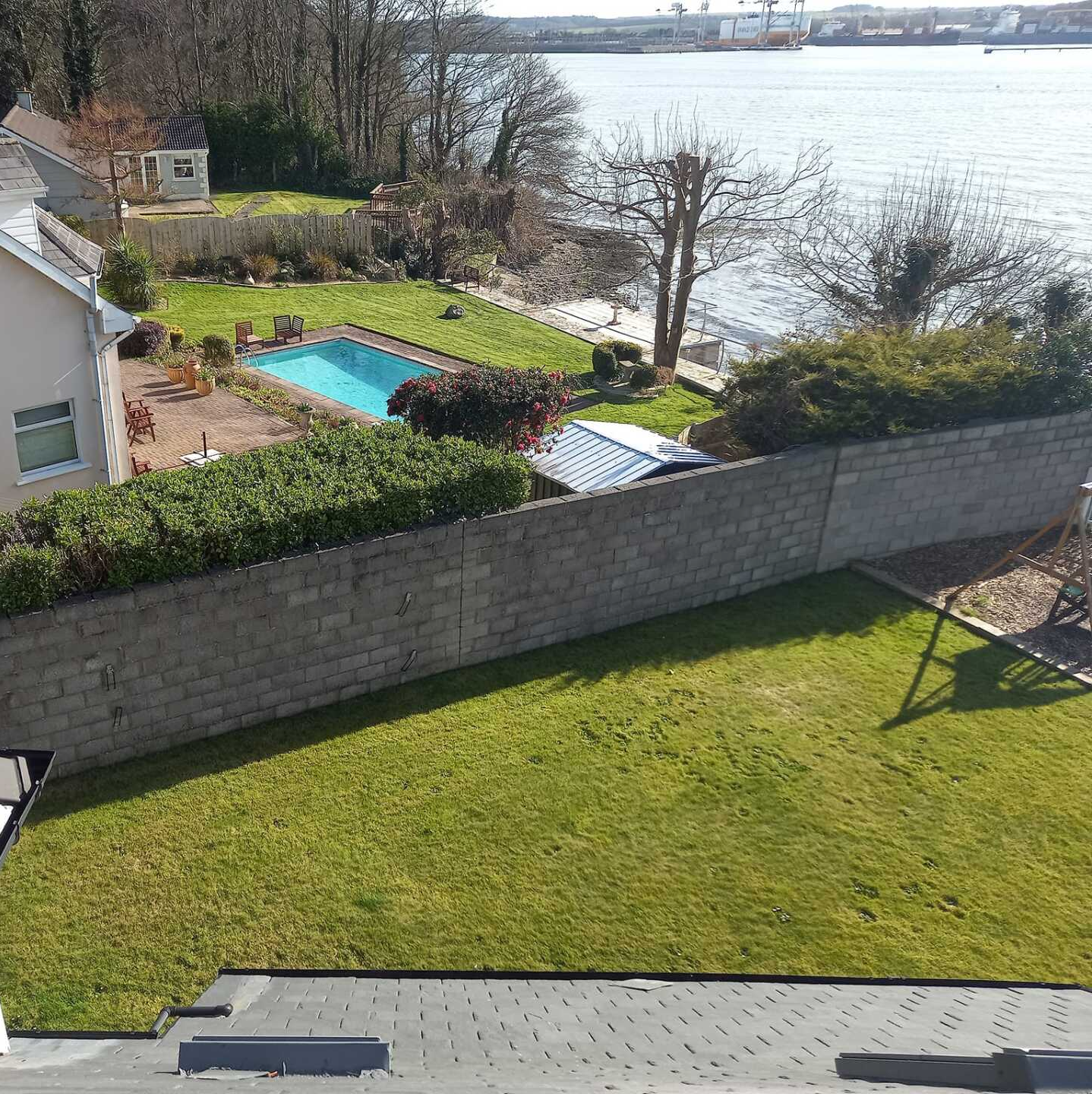 Overhead view of a house with a pool, grassy lawn, and waterfront in the background on a sunny day.