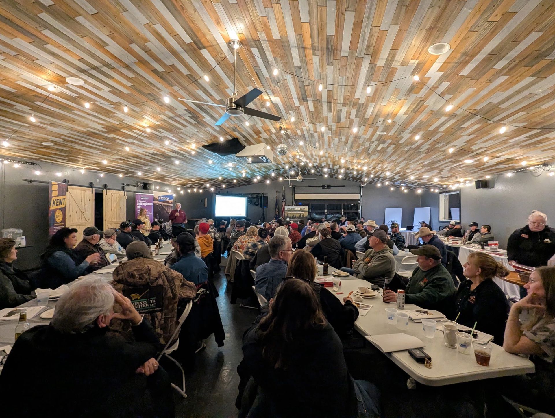Large group of people seated at long tables in a warmly lit venue; presentation on screen at the front.