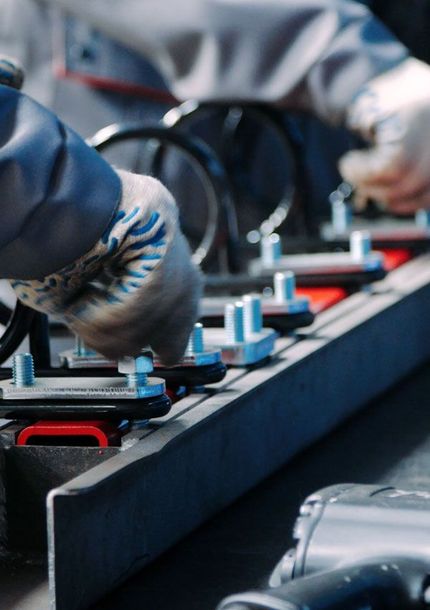Workers assembling parts on an assembly line. Hands in gloves, metallic components, and red fixtures are visible.