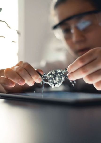 Girl with safety glasses examines a small robot spider on a tablet.