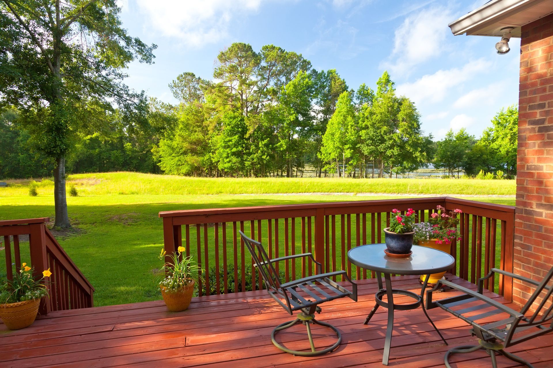 Deck builder constructed wood deck with table, chairs, railing, and steps facing open grassy field. 
