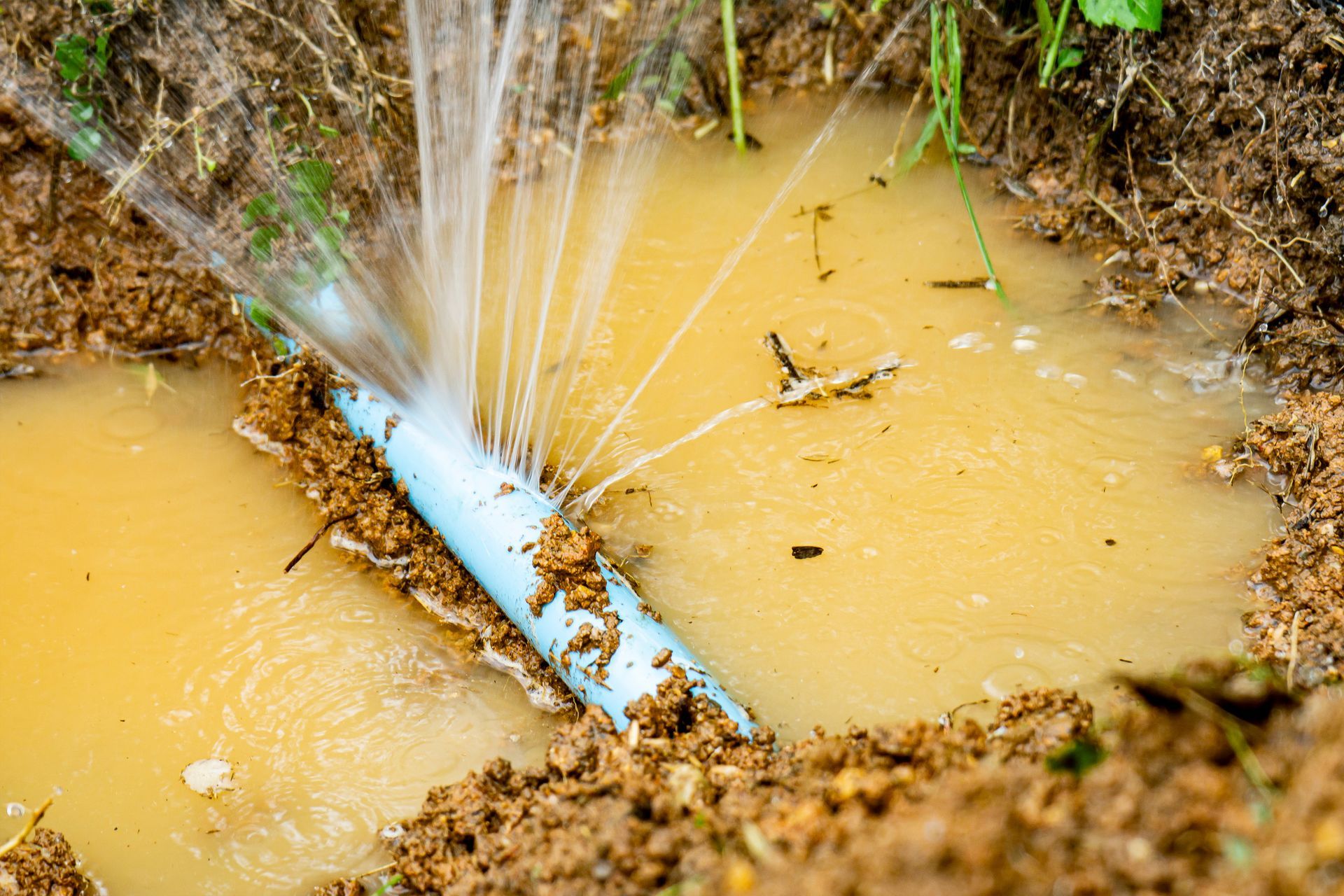 Broken blue pipe spurting water into a muddy ditch.