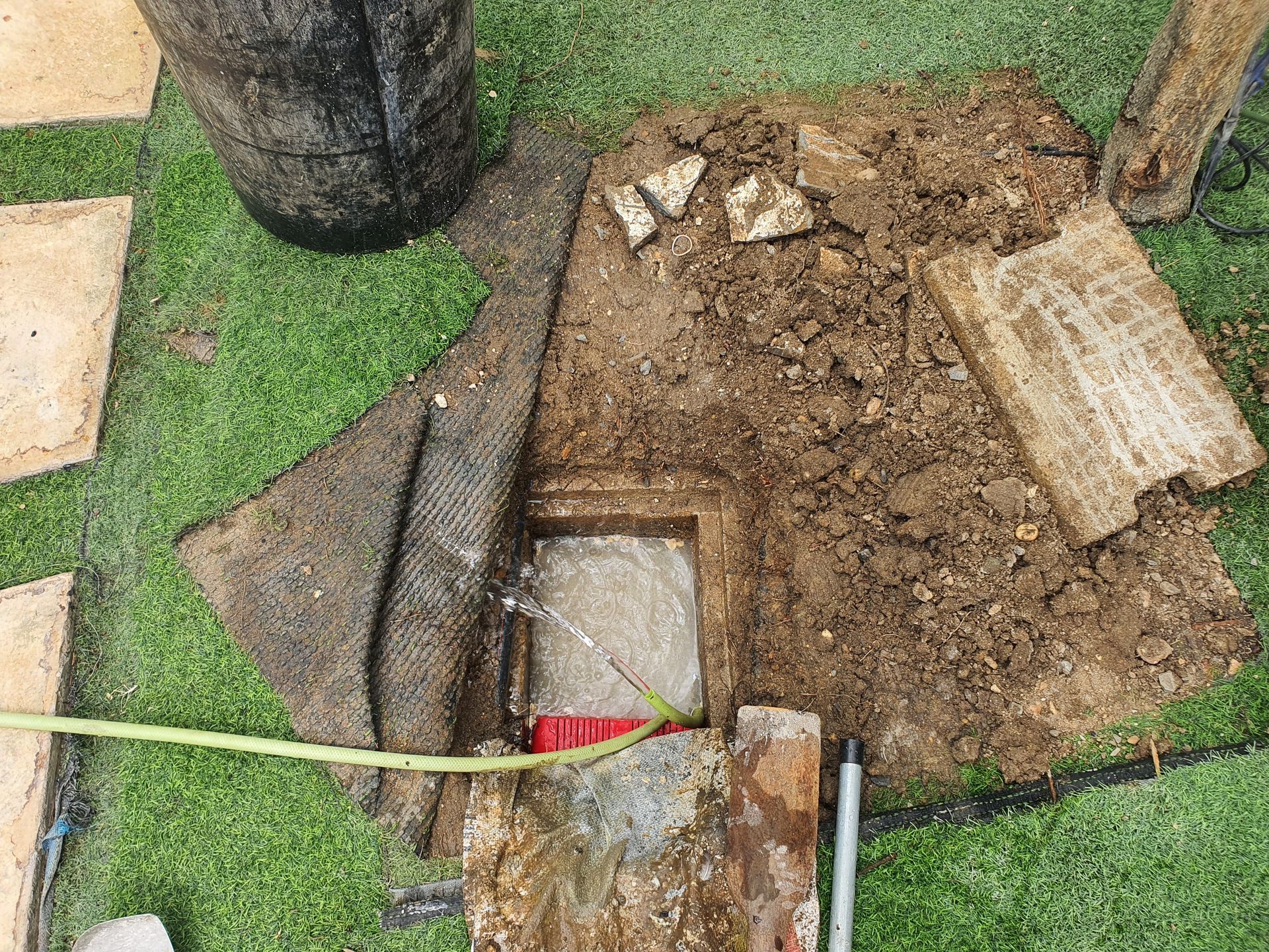 Excavated drain cover in yard. Mud, artificial turf, and a hose are visible.