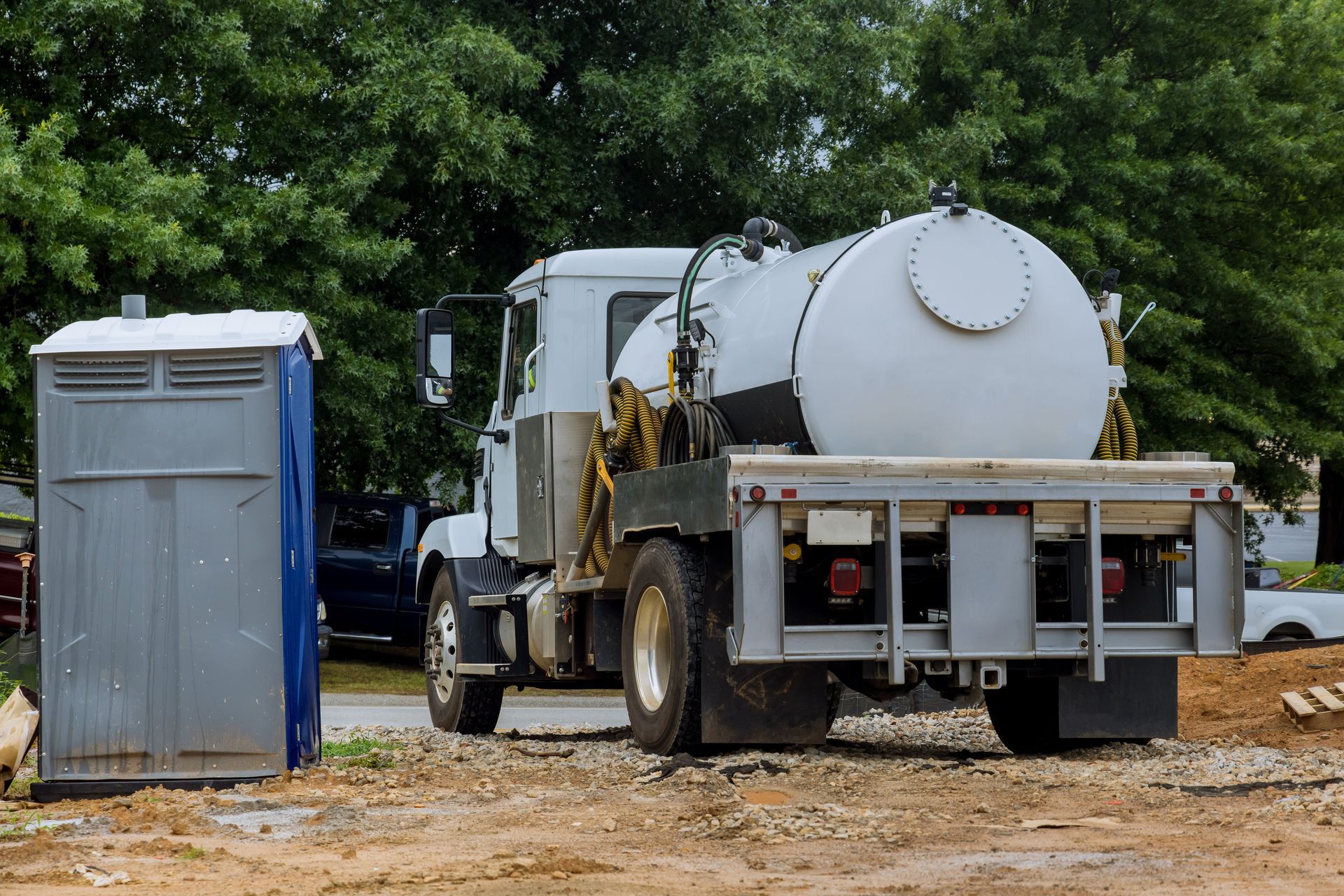 Portable toilet next to a sewage truck on a dirt road, trees in the background.