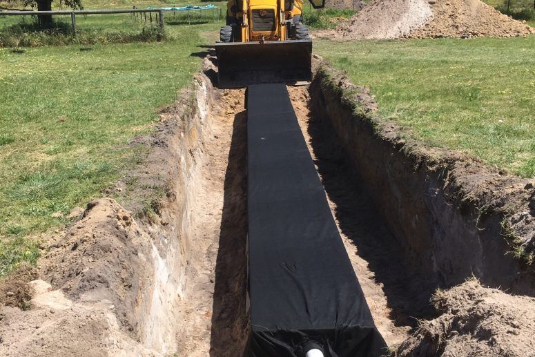 Trench with black liner being filled by a small yellow construction vehicle on a grassy field.