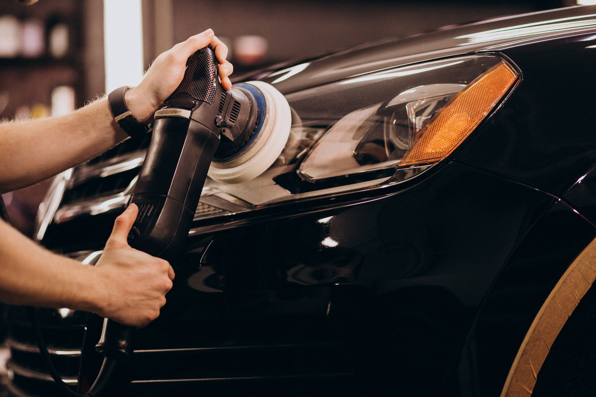 Hands polishing a black car headlight with a buffer in a workshop