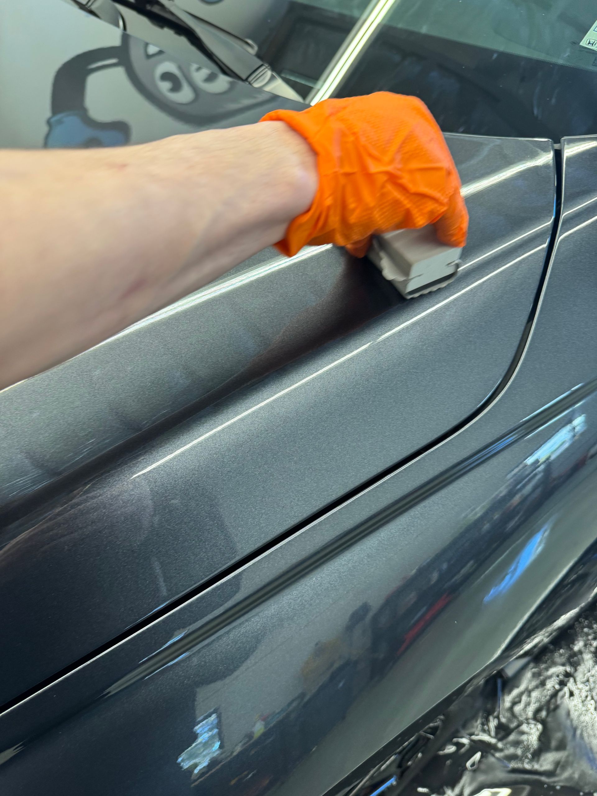 Person wearing an orange glove, applying ceramic coating to a gray car panel.