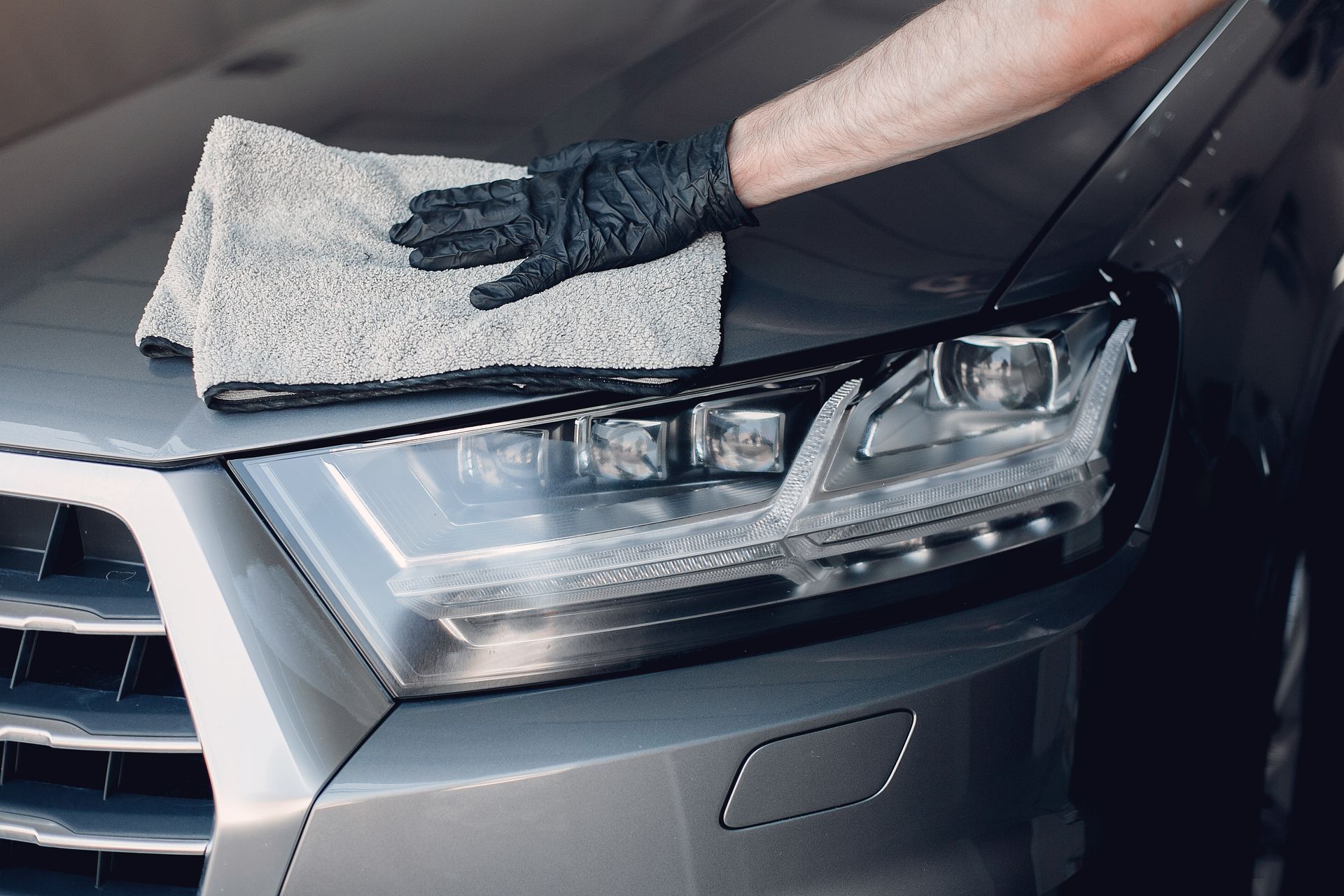 Gloved hand wiping a gray car hood with a gray microfiber cloth; cleaning the car.