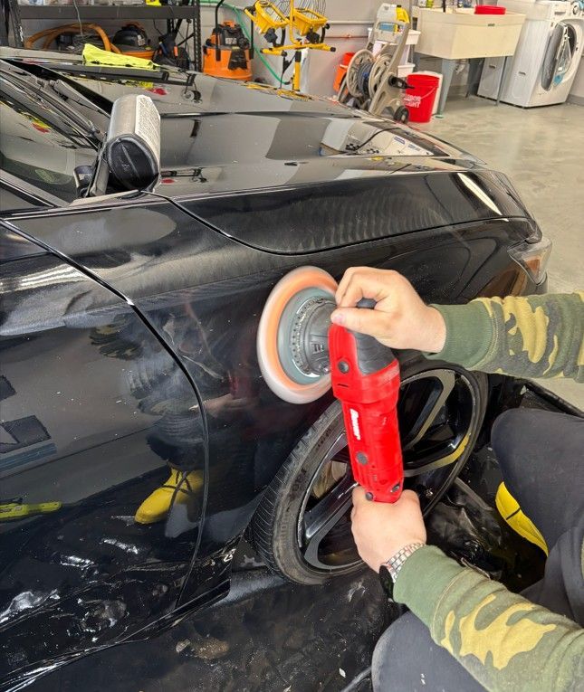 Person polishing a black car body in a workshop with a red buffer tool