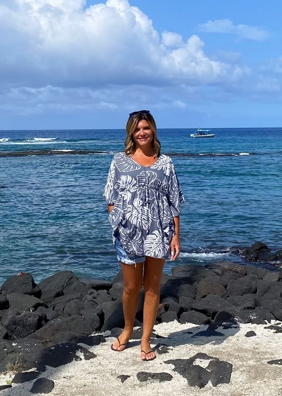 Woman standing on rocky beach in a patterned cover-up, with blue ocean and cloudy sky behind her