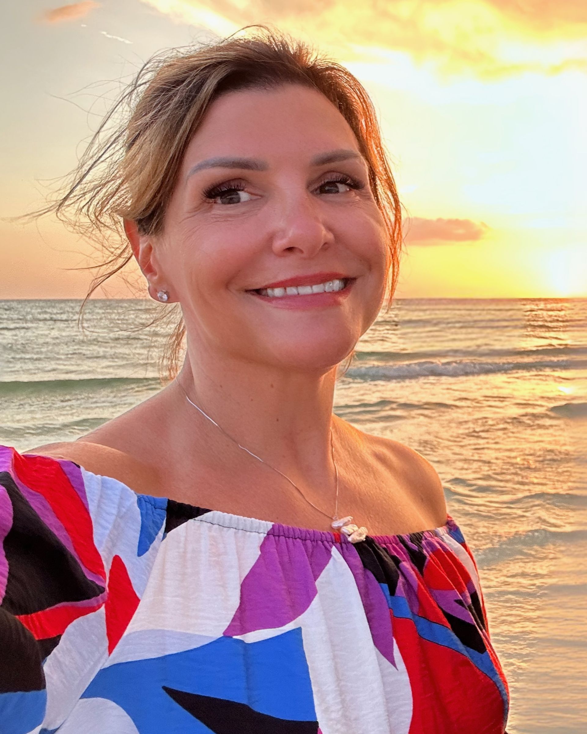 Smiling person in a colorful off-shoulder top at the beach during sunset.