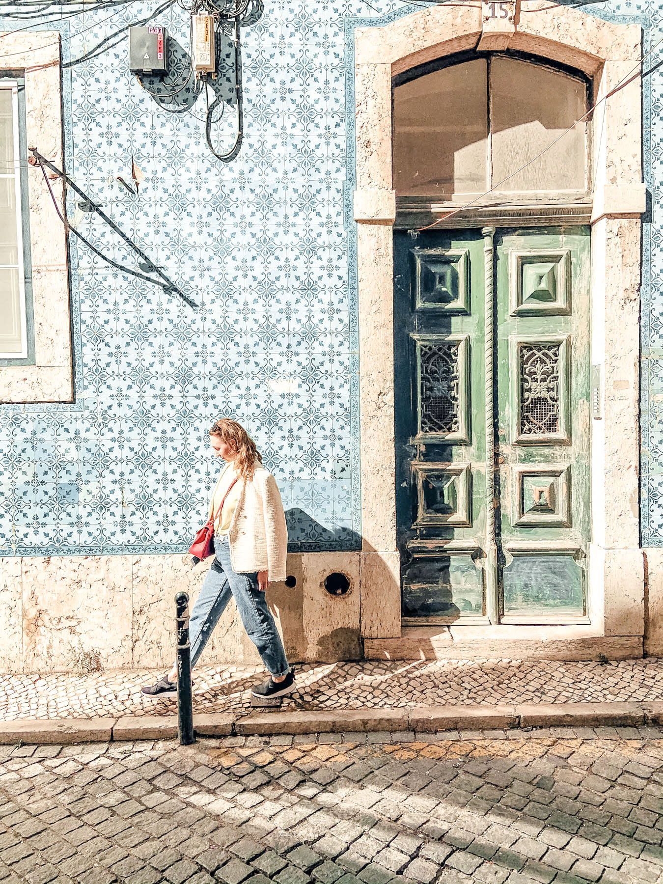 Person walking past a blue mosaic wall and green door on a cobblestone street.