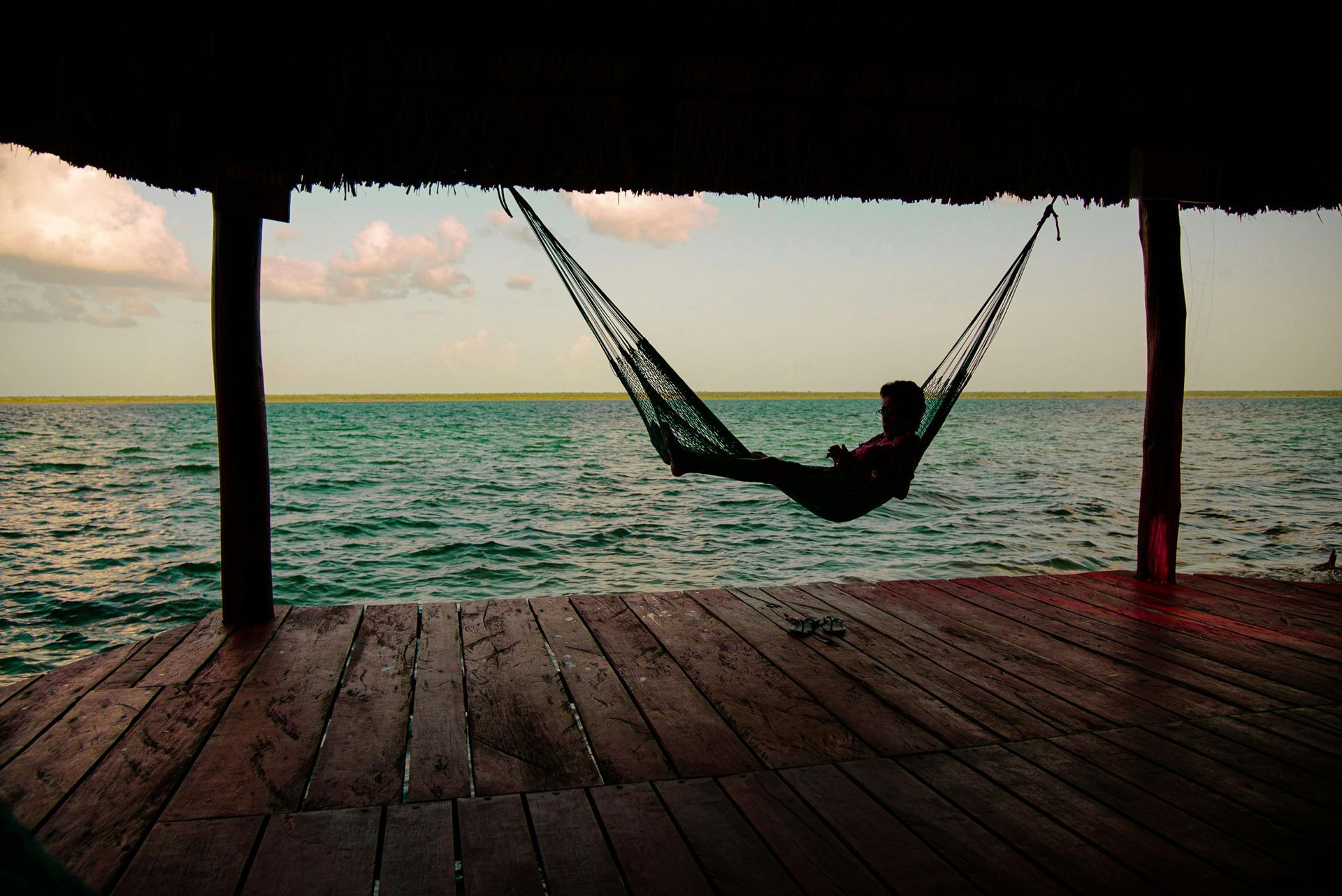 A person relaxing in a hammock suspended over a calm, blue lake inside a wooden, thatched-roof dock at sunset.