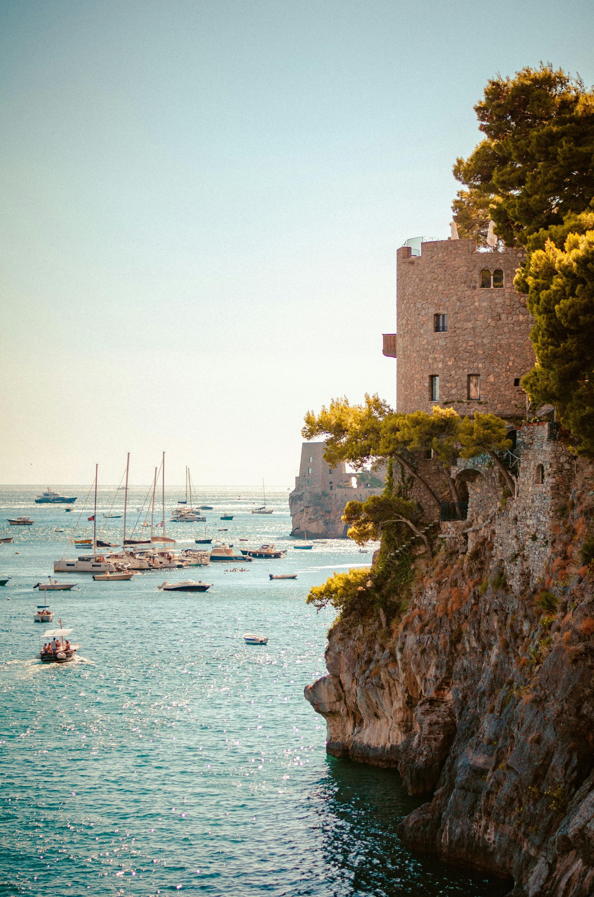 Rocky seaside cliff with a stone tower above a turquoise bay filled with small boats